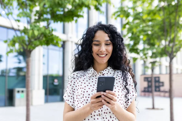 A young beautiful woman walks through the city with a phone in her hands, smiles contentedly, uses a smartphone application, browses Internet pages, types a text message, social networks.