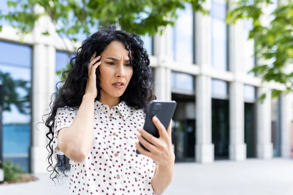 Sad disappointed woman received online notification with bad news on her phone, businesswoman walking outside office building, using application on smartphone, reading social media unsatisfied.