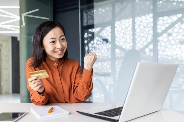 Young joyful Asian woman at workplace inside office smiling, celebrating successful online purchase in online store, businesswoman holding bank credit card in hands, using laptop.