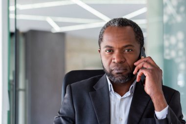 Ofisteki bir masada oturan, telefonla konuşan ve kameraya ciddi ciddi bakan kıdemli bir Afro-Amerikalı adamın yakın plan portresi..