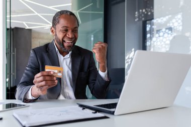 Mutlu Afrikalı genç Amerikalı adam dizüstü bilgisayarın önünde oturuyor ve mutlu, kredi kartı tutuyor ve zafer işareti yapıyor..