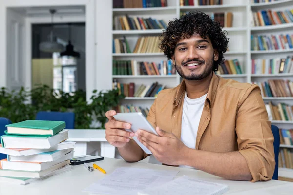Portrait of a young Indian male student sitting in the university library, holding a tablet and smiling at the camera.