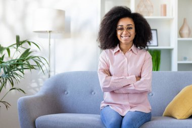 Genç ve güzel bir Afro-Amerikalı kadın pembe gömleğiyle kanepede oturuyor, kollarını göğsünün üzerinden geçirip kameraya gülümsüyor..