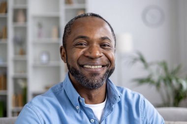 Yetişkin Afro-Amerikan bir adamın yakın plan portresi. Evdeki oturma odasında kanepede otururken gülümseyen ve kameraya bakan..
