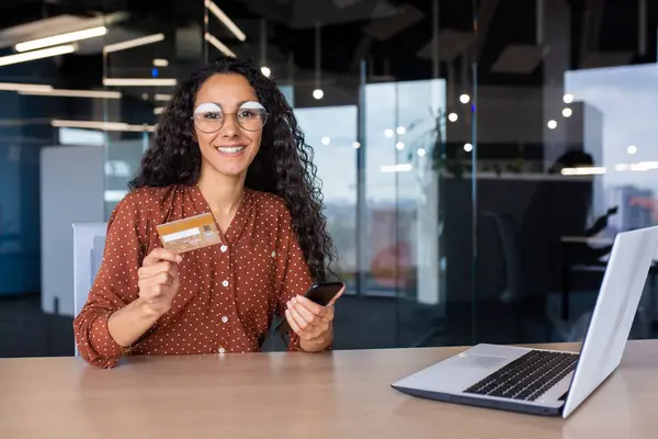 Portrait of business woman inside office at workplace, Hispanic woman smiling looking at camera, working with laptop, female worker holding phone and bank debit credit card, shopping online.