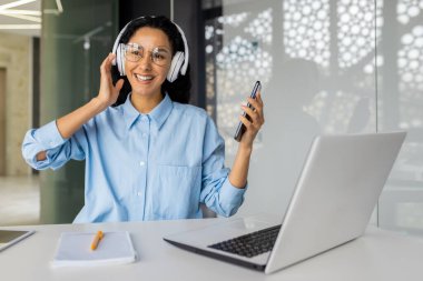 İş yerindeki genç ve güzel kadın neşeli bir şekilde müzik ve internet radyosu dinliyor, telefondaki uygulamayı kullanıyor, iş kadını molada dinleniyor, iyi iş çıkardınız..