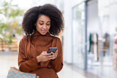 Telefon uygulaması kullanan neşeli bir kadın satın alma ve indirimleri seçiyor, alışveriş merkezi büyük, renkli alışveriş torbalarıyla geziyor..