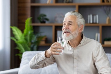 Yaşlı, gri saçlı bir adam evinde, kanepede oturur ve bir bardaktan su içer, susuzluğunu giderir, ilacını alır..