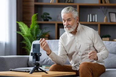 Yaşlı gri saçlı adam evdeki kanepede oturuyor ve telefonla bir video görüşmesi yapıyor, bir podcast kaydediyor, online bir konferans veriyor..