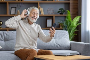 Evdeki kanepede oturan ve dinlenen kıdemli adam. Beyaz kulaklık takıyor, telefondan müzik dinliyor ve dans ediyor..