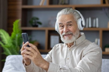 Evdeki koltukta oturan, kulaklık takan ve telefonla konuşan, gülümseyen ve kameraya bakan yaşlı, kır saçlı bir adamın yakın plan portresi..