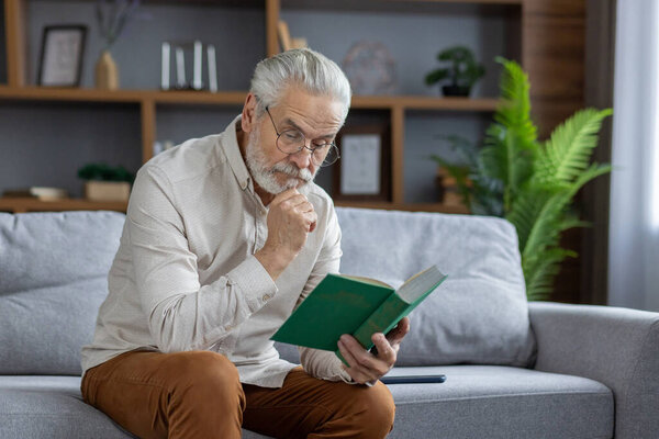 An elderly gray-haired man is sitting on the couch at home in an apron and is reading an interesting book with concentration.