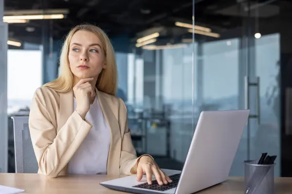 Serious concentrated thinking woman at workplace inside office working with laptop, businesswoman brainstorming strategy plans financial tasks.
