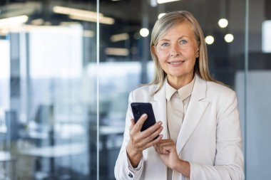 Ofiste takım elbiseli gülümseyen gri saçlı bir iş kadınının portresi. Telefonu elinde tutuyor ve kameraya güvenle bakıyor..