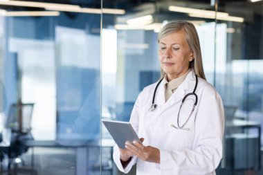 Klinikte durup tablet kullanan ciddi bir yaşlı, gri saçlı kadın doktor. Yakın plan fotoğraf..