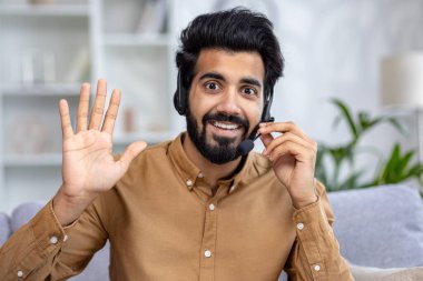 Kamera görüntüsü: Kablosuz kulaklıklı, pozitif gülümseyen Hintli adam uzaktan video görüşmesi yaparken, açık evdeki kanepede oturan online konferans röportajı.