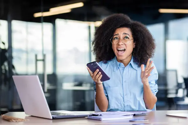 Upset frustrated and sad businesswoman, got bad news online on phone, female employee reading news nervously, working inside office with laptop, woman with curly hair depressed.