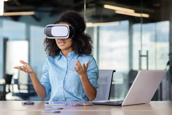 Businesswoman at workplace wearing virtual reality glasses communicating remotely in video simulator, female office worker working with laptop, remote meeting.