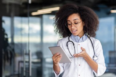 Genç bir kadın doktor elinde bir tablet bilgisayar tutuyor, beyaz önlüklü bir kadın hastalara uzaktan danışıyor, tatmin olmuş bir şekilde gülümsüyor, pencerenin yanında duruyor..
