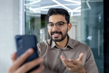 İş yerindeki başarılı genç işadamı telefondaki uygulamayı kullanarak video görüşmesinde konuşuyor, işyerinde gülümseyen adamı kapatıyor..