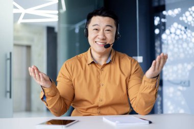 Ofiste oturan gülümseyen Asyalı genç bir adamın portresi. Bir kamera önünde, kulaklıkla. Ellerini açıp kameraya gülümsüyor..