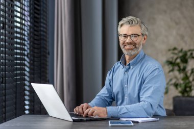 Olgun gri saçlı iş adamı portresi, kıdemli deneyimli bir adam gülümseyen ve kameraya bakan, penceresi pencereli ve ofis içinde panjurları olan masa başında oturan.