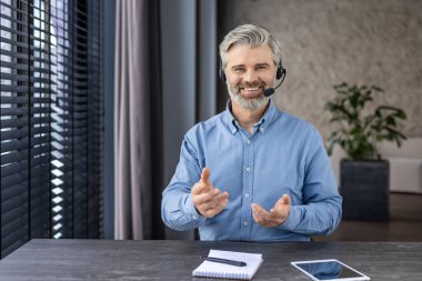 Webcam görüntüsü, olgun, gri saçlı patron iş yerindeki kulaklıkla kameraya bakıyor. İnternetteki toplantıda gülümseyen adam, tişörtlü kıdemli iş adamı..