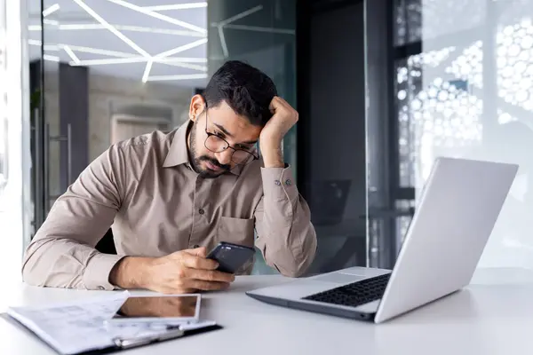 Upset sad man at workplace inside office, businessman uses phone reads bad news received by e-mail, employee disappointed holds smartphone in hands browses internet sites and social networks.