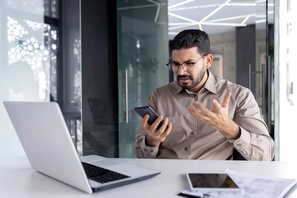 Upset sad man at workplace inside office, businessman uses phone reads bad news received by e-mail, employee disappointed holds smartphone in hands browses internet sites and social networks.