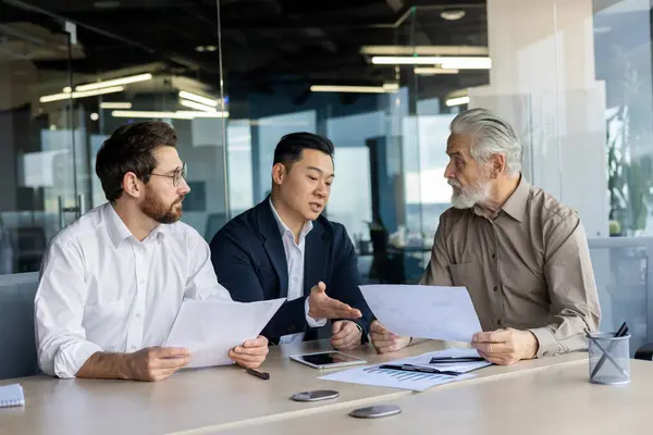 Three diverse business people talking together sitting at a meeting desk inside office, businessmen in business suits discussing with boss business owner about financial indicators, achievements.