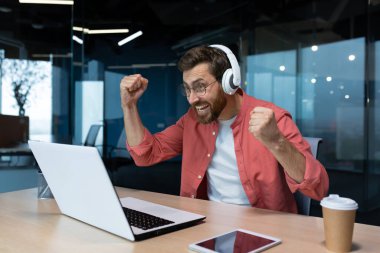 Ofisteki işyerindeki adam, bilgisayardaki bir spor maçını izlerken tezahürat yapıyor, kulaklıklarla seviniyor, spor takımının sonucundan memnun..
