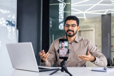 Başarılı bir işletme eğitmeni olan öğretmen çevrimiçi eğitim videosu kursunu kaydediyor iş yerinde telefon ve tripodu kullanan bir adam dizüstü bilgisayarla masada oturuyor..