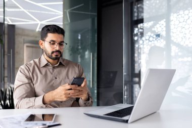 İş yerindeki ciddi konsantre işadamı, elinde bir akıllı telefon tutuyor, dizüstü bilgisayarı olan bir işçi interneti ve sosyal ağları tarıyor, mesaj yazıyor..