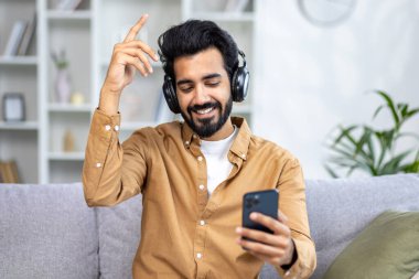 Genç Hintli bir adam evinde, kanepede kulaklıkla oturur, telefondan müzik dinler, gözlerini kapatır, elleri havada dans eder, dinlenir..