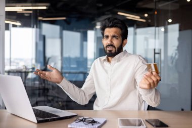Kredi kartı sorunları. Ofisteki masada oturan genç Hintli bir adamın portresi. Endişeli bir şekilde kameraya bakıyor ve hayal kırıklığı içinde ellerini kaldırıyor..