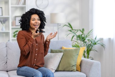 Genç bir Afro-Amerikalı kadın evdeki koltukta oturuyor ve hoparlörden cep telefonuyla konuşuyor..