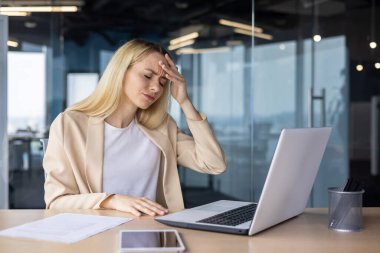 Yorgun ve çok çalışan genç Ukraynalı kadın laptopun ofisinde çalışıyor, başını tutuyor, şiddetli bir acı ve baskı hissediyor..