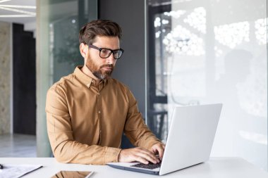 Ofisin içinde dizüstü bilgisayarla çalışan ciddi odaklı iş adamı gömlekli ve gözlüklü olgun bir adam klavyede yazan, programcı..