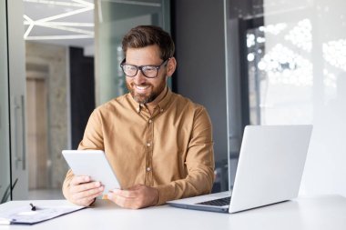 Genç bir erkek programcı dizüstü bilgisayarın masasında çalışıyor ve elinde bir tablet tutuyor..