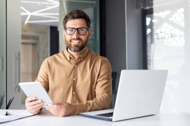 Bir ofis merkezinde çalışan genç bir işadamının portresi, bir masada dizüstü bilgisayarla kitap okuyor ve elinde tablet tutuyor, gülümsüyor ve kameraya bakıyor..