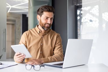 Genç bir erkek programcı ve geliştirici masadaki ofiste oturuyor ve bir dizüstü bilgisayar ve tabletle çalışıyor. Görüntülü arama üzerine konuşur.
