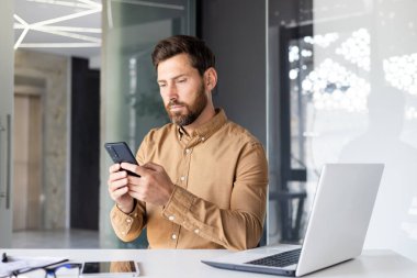 Ofiste çalışan genç bir işadamı, elinde laptop ve belgelerle bir masada oturuyor ve cep telefonu kullanıyor..