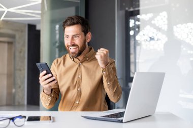 Kazanan adam telefonu elinde tutuyor, işyerinde çalışan işadamı online başarı sonuçlarından memnun, çalışan çalışan akıllı telefon uygulaması kullanıyor, ofiste dizüstü bilgisayarla oturuyor, günlük gömlekler giyiyor..
