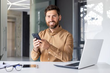 Ofis içinde başarılı bir iş adamının portresi, kameraya gülümseyen bir erkek, cep telefonlu bir işçi, online uygulama kullanıyor..