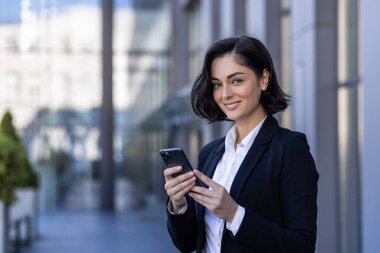 Ofis binasının yanında elinde telefonla kameraya gülümseyen genç bir iş kadını portresi..