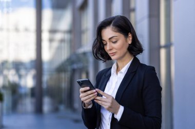 Genç bir iş kadını iş elbisesi içinde ofis merkezinin yanında duruyor ve telefonu kullanıyor, mesajlar yazıyor, mesajlar yazıyor..