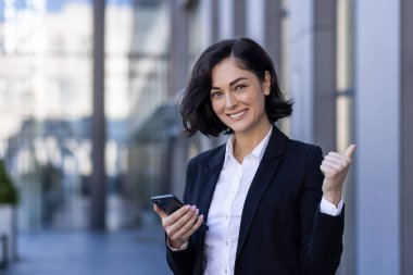 Bir ofis merkezinin yanında duran, elinde telefonla kameraya gülümseyen başarılı genç bir iş kadınının portresi..