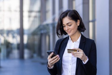 Genç gülümseyen iş kadını dışarıda takım elbisesiyle duruyor ve kredi kartı ve telefon kullanıyor..