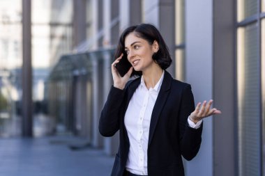 Genç başarılı iş kadını telefonda konuşuyor. İş elbisesi giymiş bir kadın. Ofis binasının dışında dolanıyor..