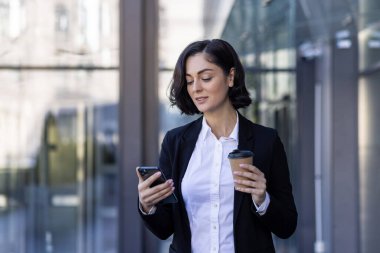 Genç bir kadın ofis çalışanı, iş molasında, elinde kahve ve telefonu kullanarak bir büro merkezinin önünde duruyor..